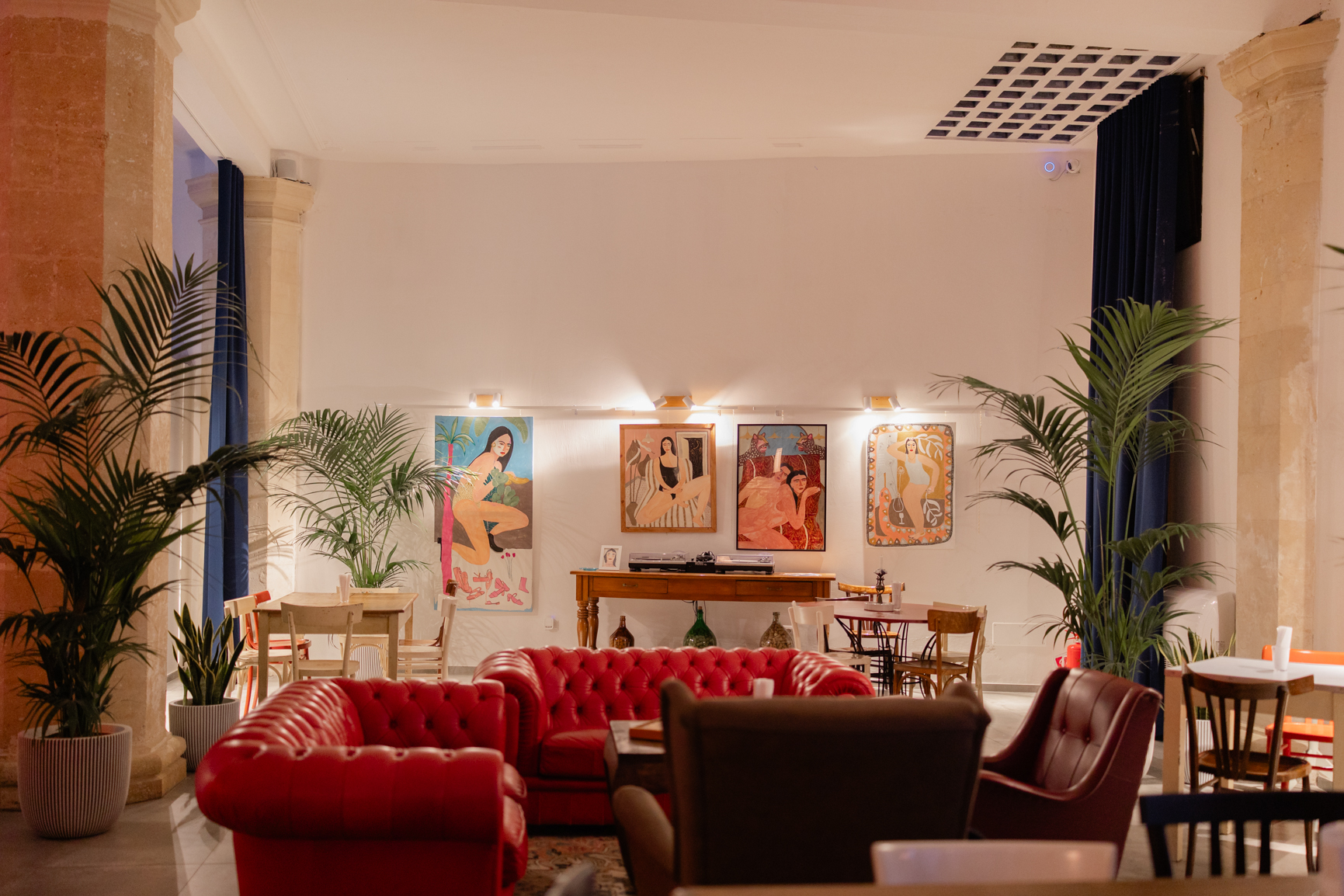 La sala di Vineria La Mara: divani Chesterfield rossi fra le colonne in pietra leccese di Palazzo Andretta, tende blu, palme e quadri di artisti locali illuminati alle pareti